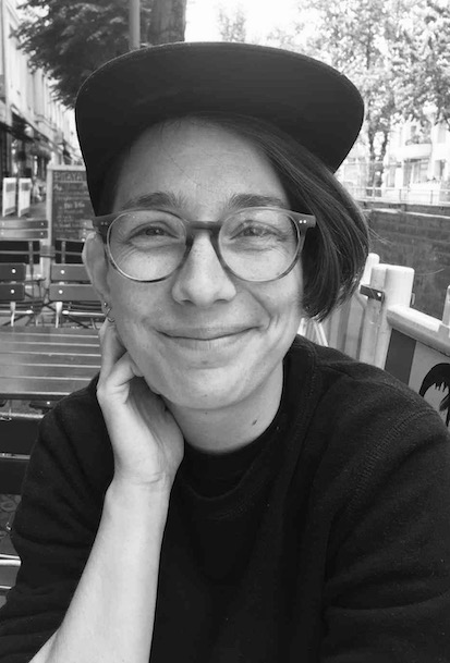 Young white woman sitting at an outdoor cafe, wearing glasses, a black sweater and a black baseball cap.